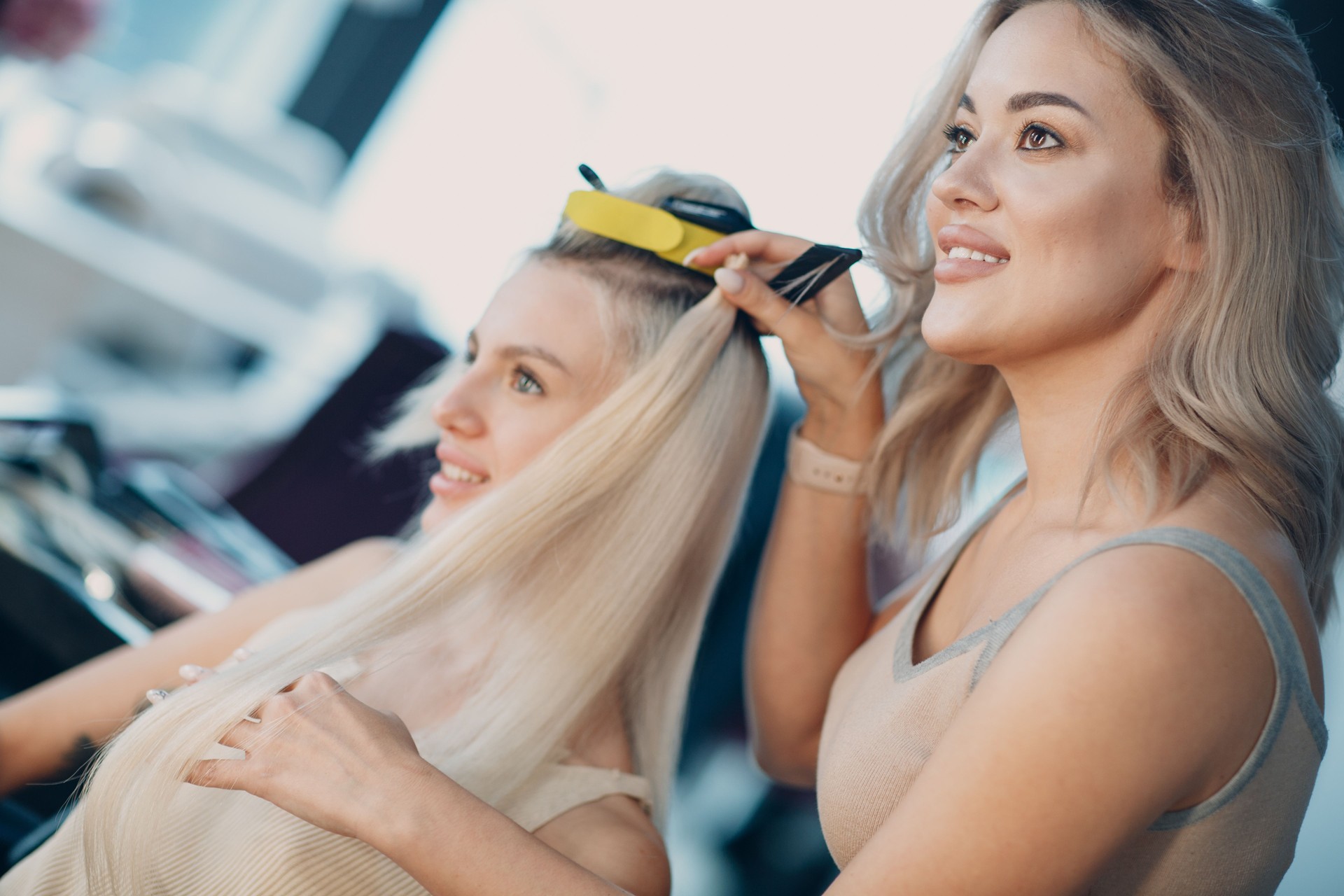 Hairdresser female making hair extensions to young woman with blonde hair in beauty salon. Professional strand hair extension.